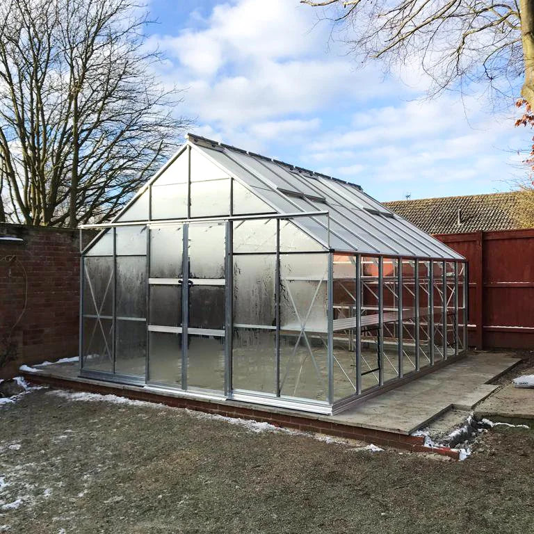 A large glass Rhino Greenhouse stands on a raised concrete platform surrounded by a fenced garden; leafless trees are visible under a partly cloudy sky.
