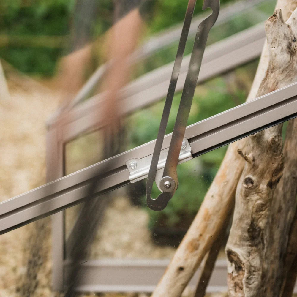 Metal Rhino Greenhouse window actuator opens a glass panel, supported by a sturdy frame. Wooden beams and blurred greenery are visible in the background, suggesting a garden setting.