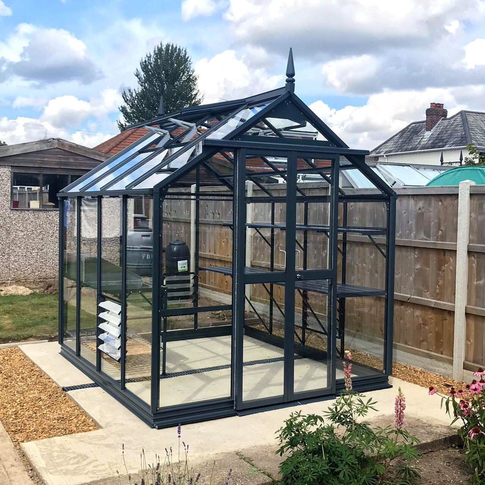 Rhino Greenhouse stands on a concrete slab, surrounded by a garden with various plants. It features glass panels and a pointed roof, enclosed by a wooden fence under a partly cloudy sky.