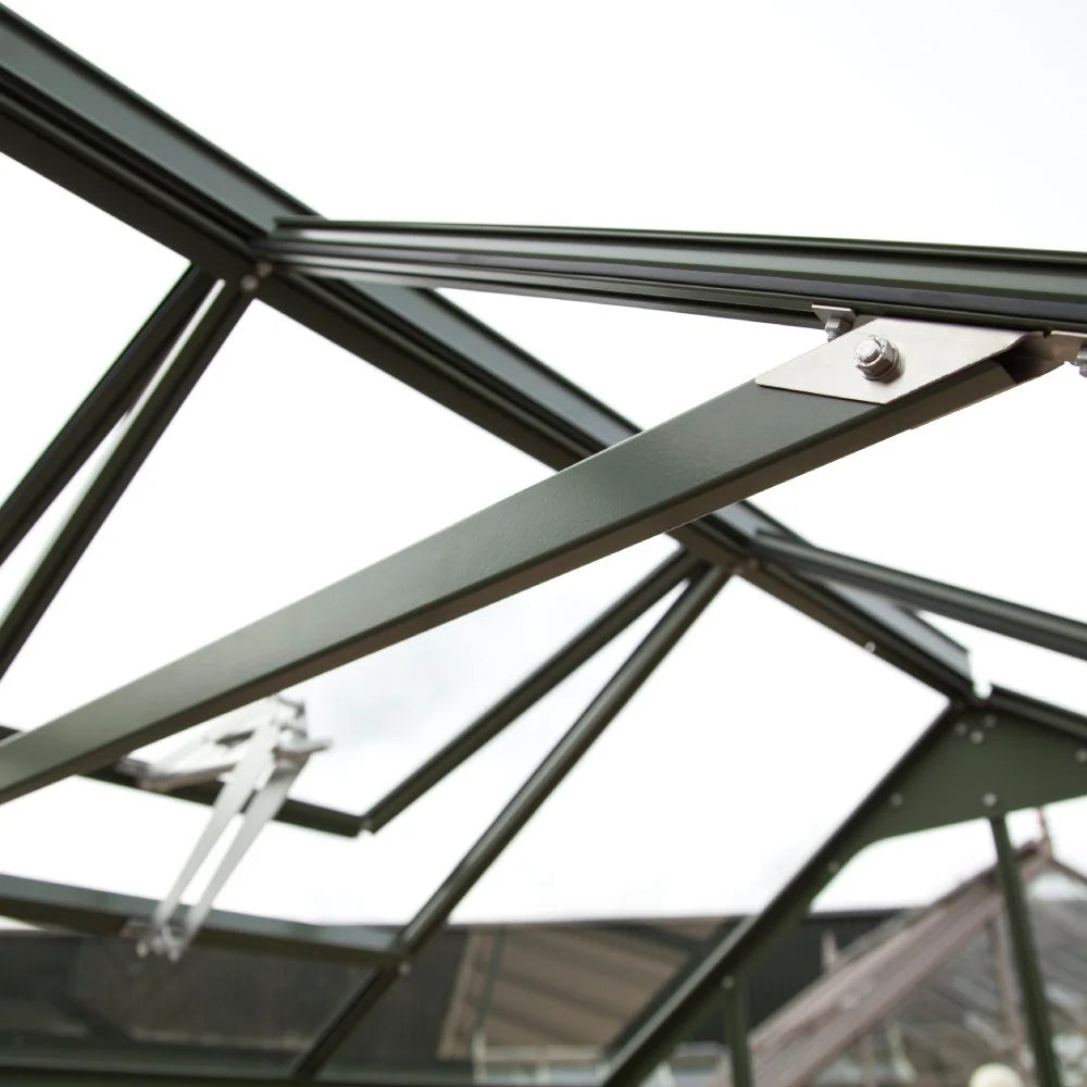 A dark green metal Rhino Greenhouse frame is shown from below, with connected beams and bolts, set against an overcast sky.