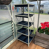 A metal shelving unit with black trays stands in a Rhino Greenhouse on a wooden floor. Nearby, there's a planter with red flowers, and outside, blurred figures and more Rhino Greenhouses are visible.