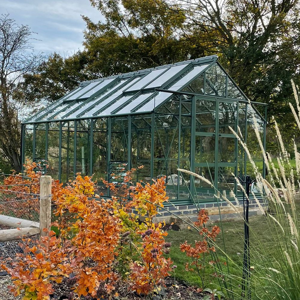 A green glass Rhino Greenhouse stands amid an autumnal garden, surrounded by colorful trees and foliage under a clear sky.