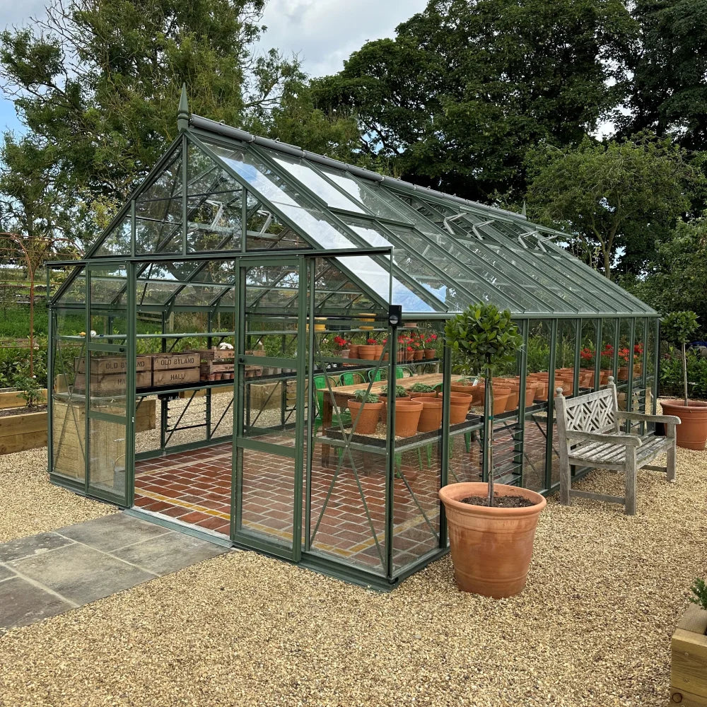 A glass Rhino Greenhouse standing on a gravel surface contains potted plants on shelves, with a wooden bench nearby, surrounded by trees and greenery.