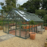 A glass Rhino Greenhouse standing on a gravel surface contains potted plants on shelves, with a wooden bench nearby, surrounded by trees and greenery.