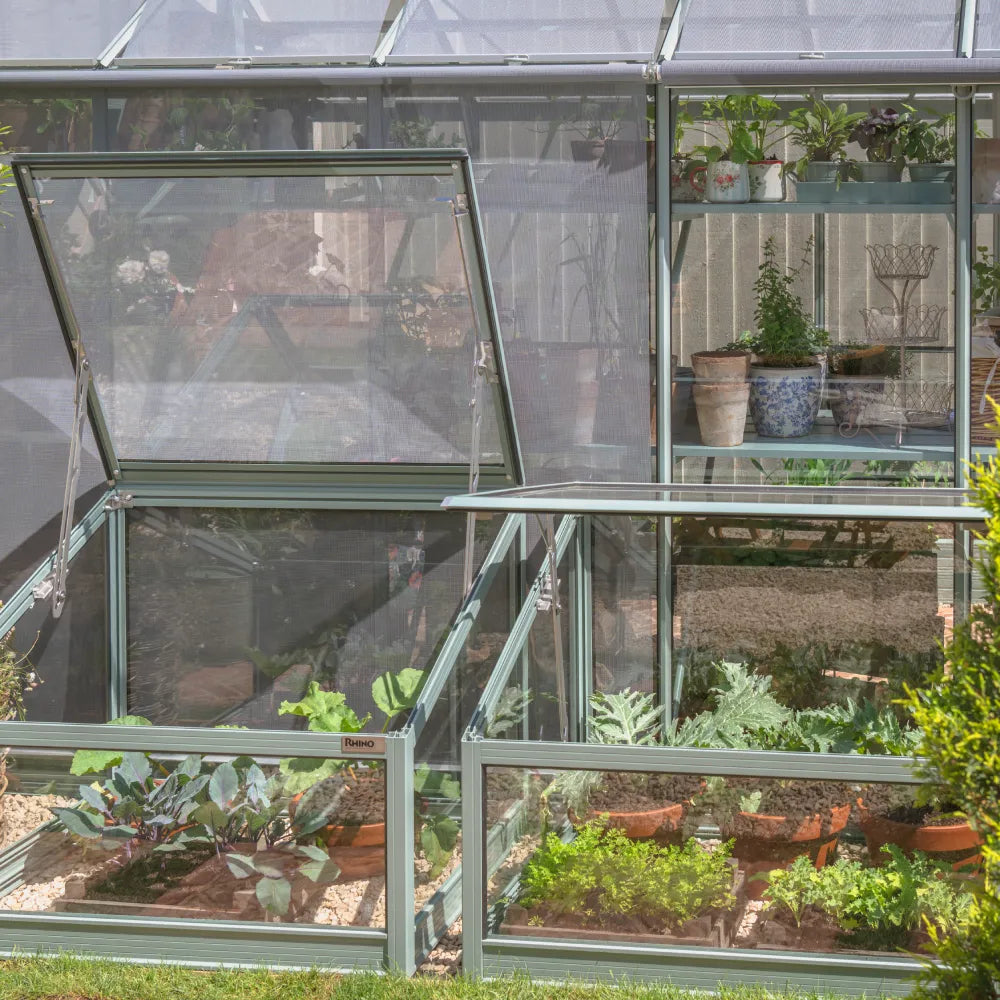 Rhino Greenhouse with open panels housing various potted plants and greenery, situated on gravel. Label reads Rhino on a frame, surrounded by a garden environment.