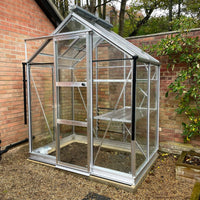 A small glass Rhino Greenhouse stands on gravel, attached to a brick wall, surrounded by greenery. It has a metal frame and a partially open door.