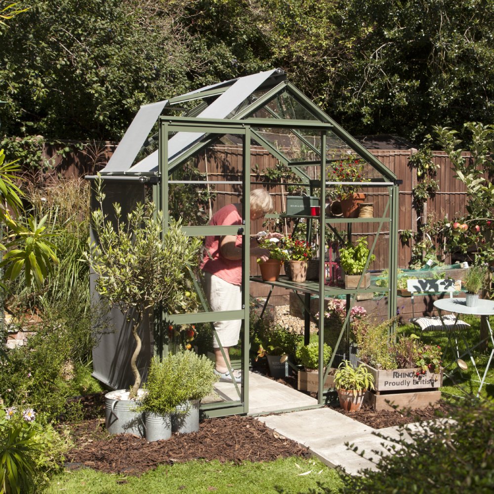 A person tends to plants inside a green metal and glass Rhino Greenhouse with open roof panels. The garden is lush with potted plants, and Rhinogreenhouses boxes are visible nearby.