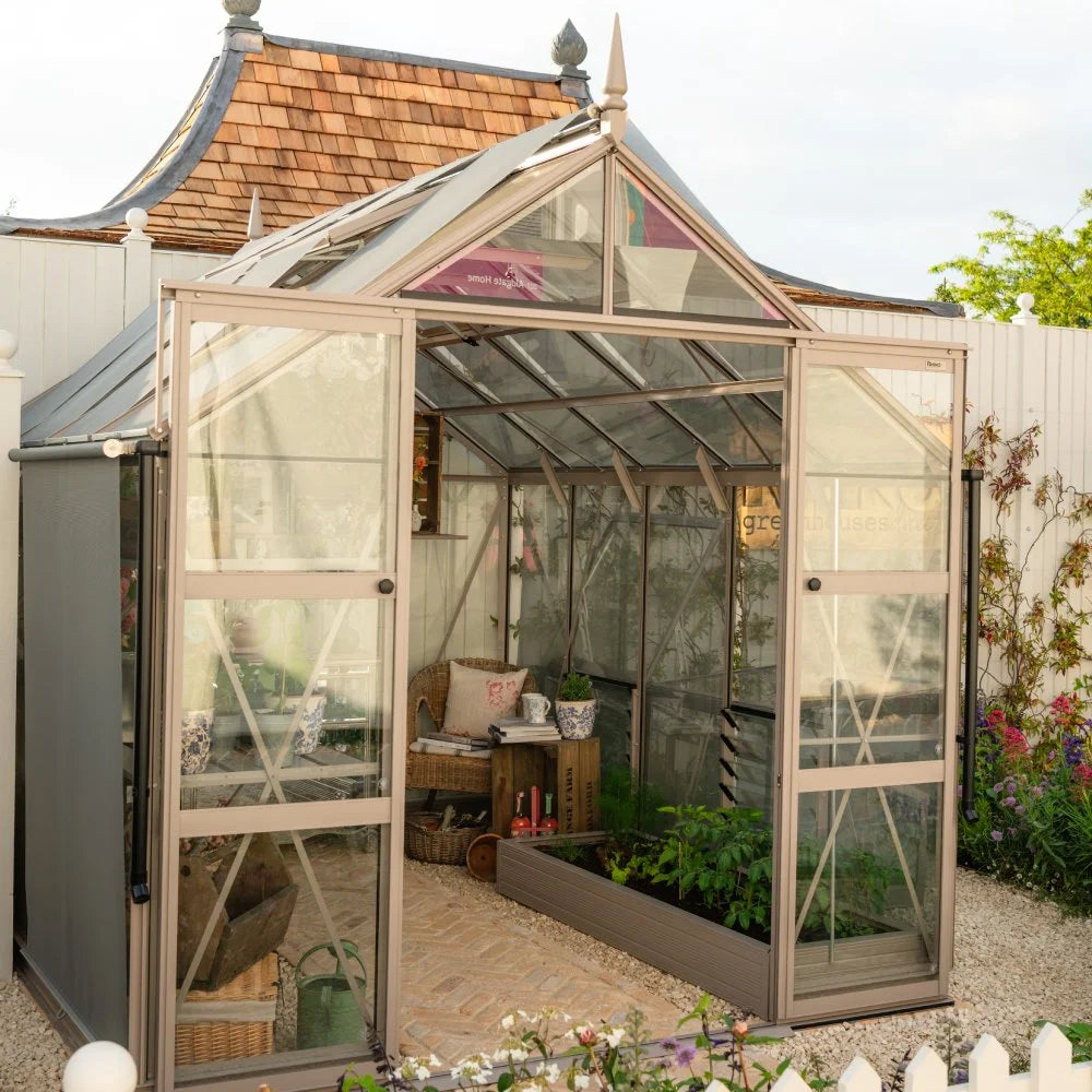 A Rhino Greenhouse with transparent panels displays plants and gardening tools on a brick floor. It is situated in a fenced garden area, surrounded by various flowers and greenery.