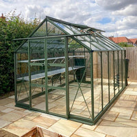 A green-framed glass Rhino Greenhouse, with open roof vents, stands on a stone patio in a garden surrounded by wooden fences and lush greenery under a cloudy sky.