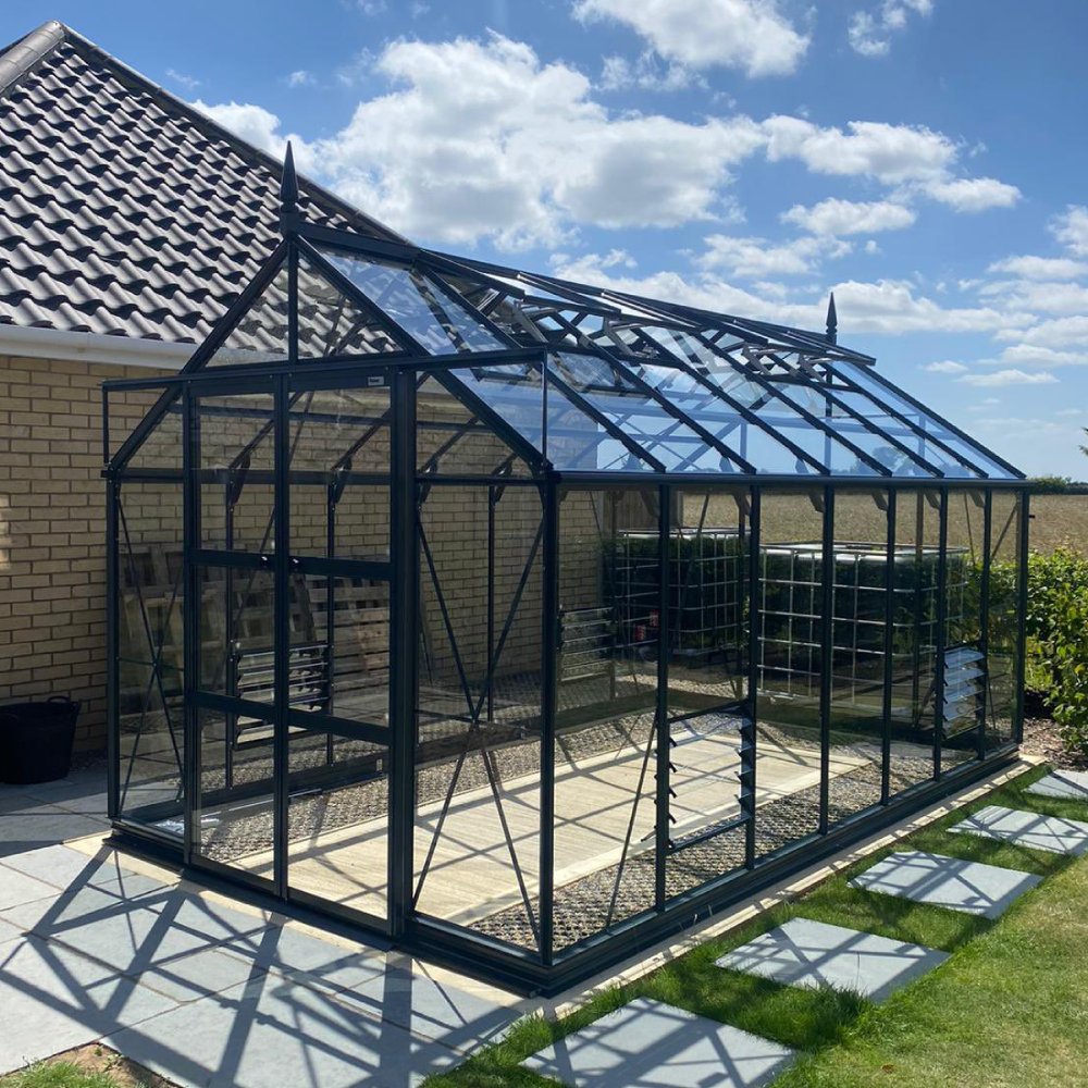 A large glass Rhino Greenhouse stands attached to a brick house, reflecting sunlight. It is surrounded by a neatly landscaped garden with paving stones and a clear blue sky overhead.