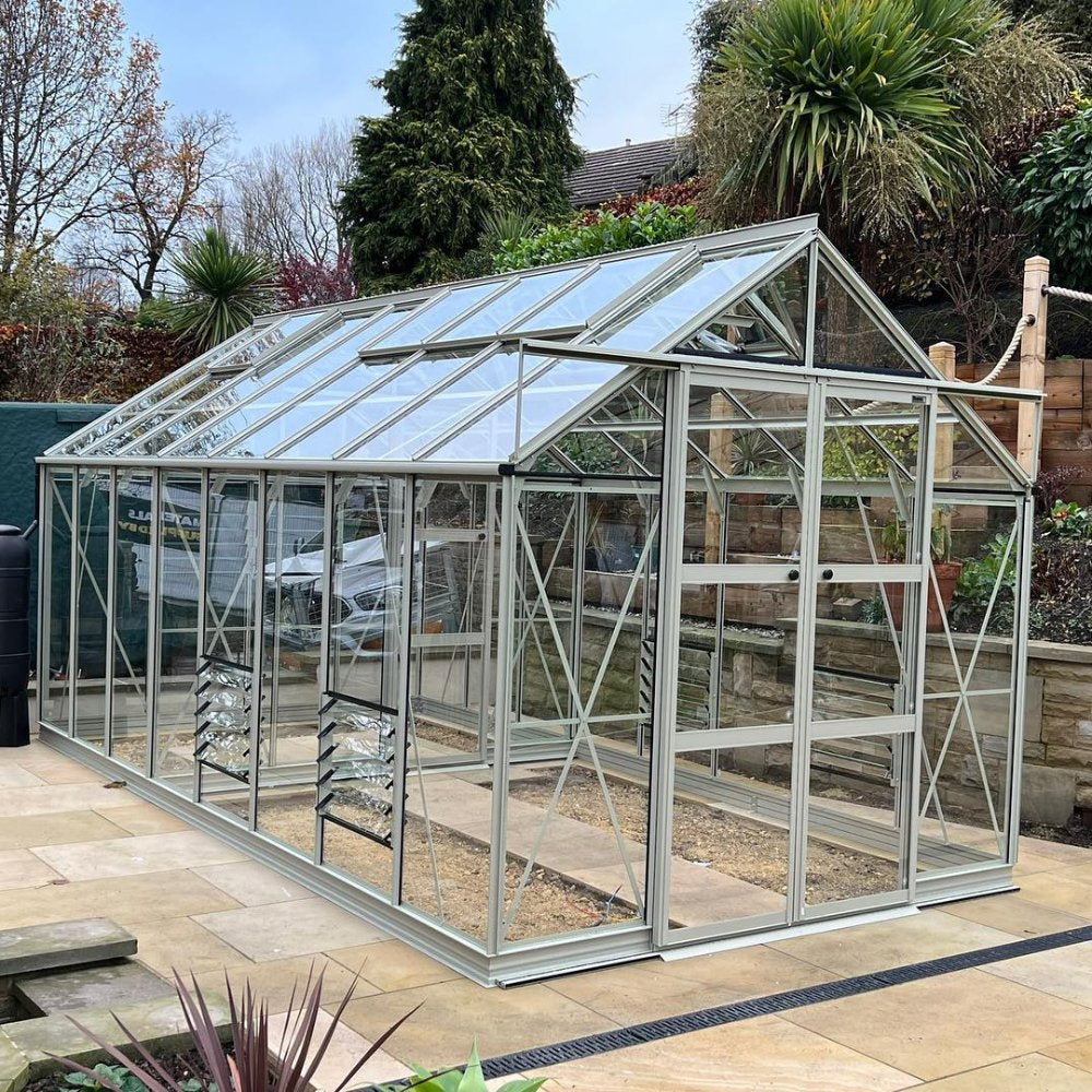 A large, glass-paneled Rhino Greenhouse with an aluminum frame stands on a paved patio, surrounded by lush greenery and stone walls, under a clear blue sky.