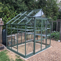 A green, glass Rhino Greenhouse stands on a gravel surface with open shelves inside. It's surrounded by leafy bushes and wooden fencing in a garden setting.