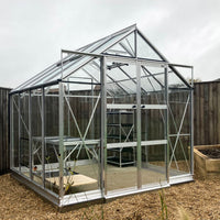 A glass Rhino Greenhouse with a metal frame stands on a gravel surface, featuring shelving inside and surrounded by a wooden fence and a planter in a garden setting.