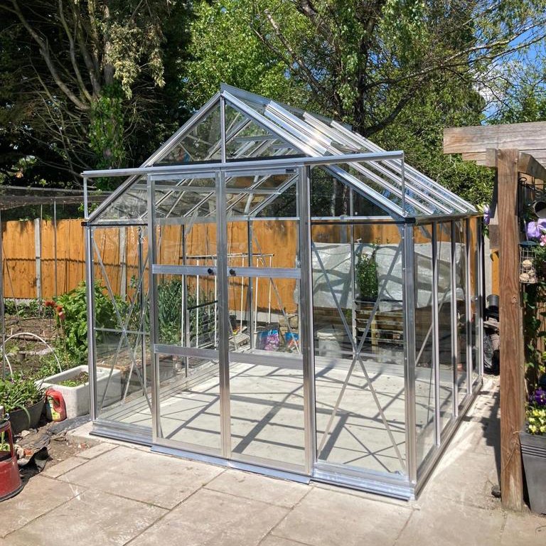 A glass and metal-framed Rhino Greenhouse stands on a patio, surrounded by garden plants and a wooden fence, under a clear blue sky with trees in the background.