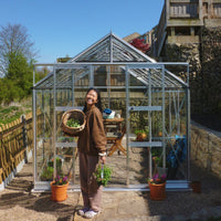 A person holds a basket of vegetables and plants, smiling in front of a glass Rhino Greenhouse surrounded by potted plants and wooden fencing, under a clear blue sky.