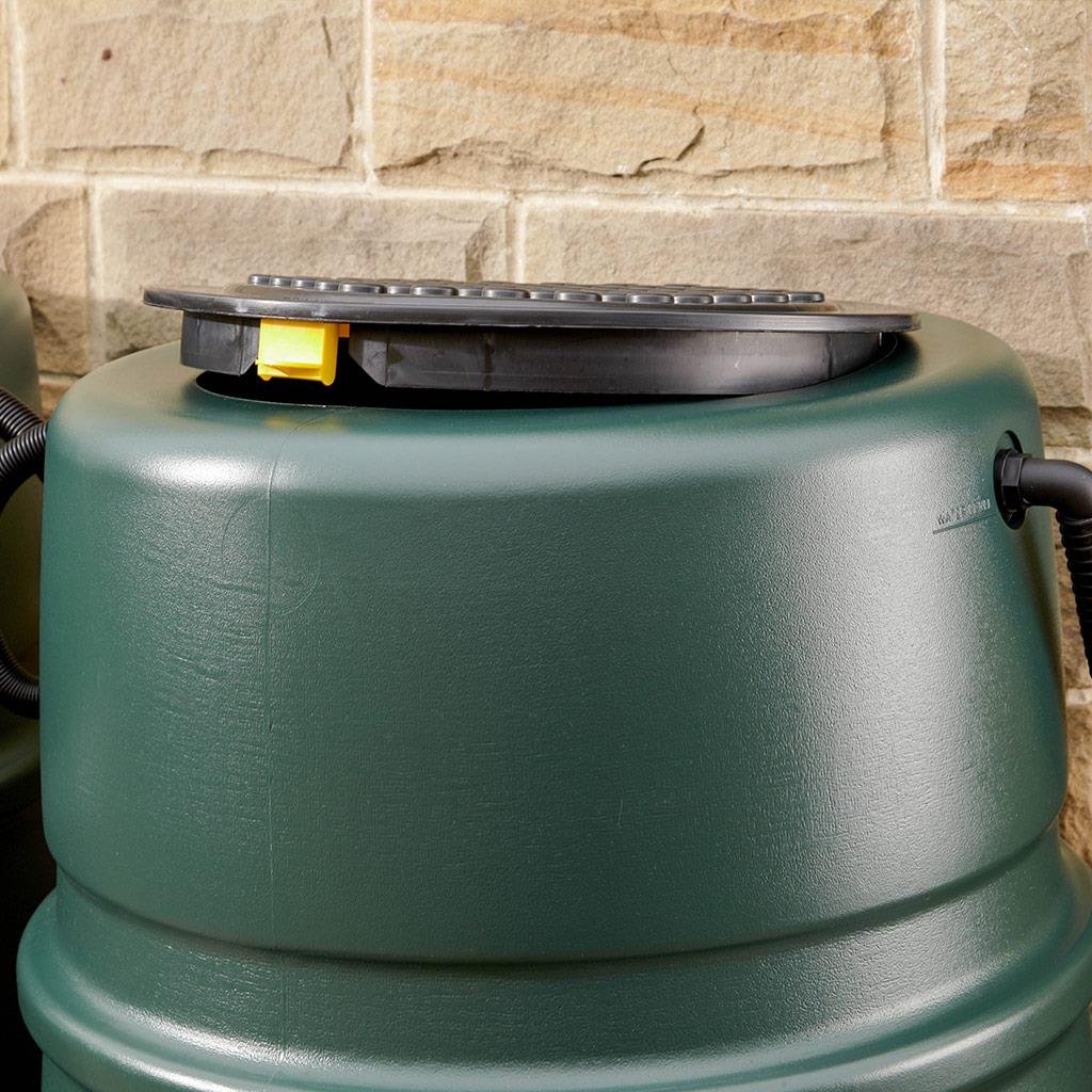 A green water butt with a black lid and yellow handle stands against a stone wall, featuring a hose connector on the side.