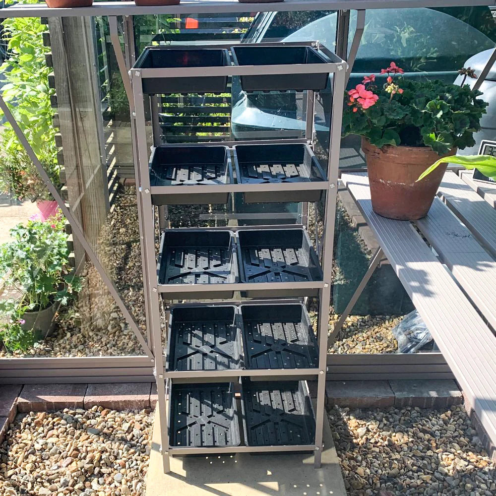 A tiered metal shelf holds black seed trays, positioned inside a glass Rhino Greenhouse. Nearby, potted flowers and plants sit on a wooden bench, surrounded by pebbled ground.