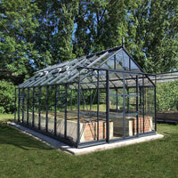 A glass Rhino Greenhouse with a dark metal frame stands on a concrete base, surrounded by grass and lush trees, allowing natural sunlight to filter through its structure.