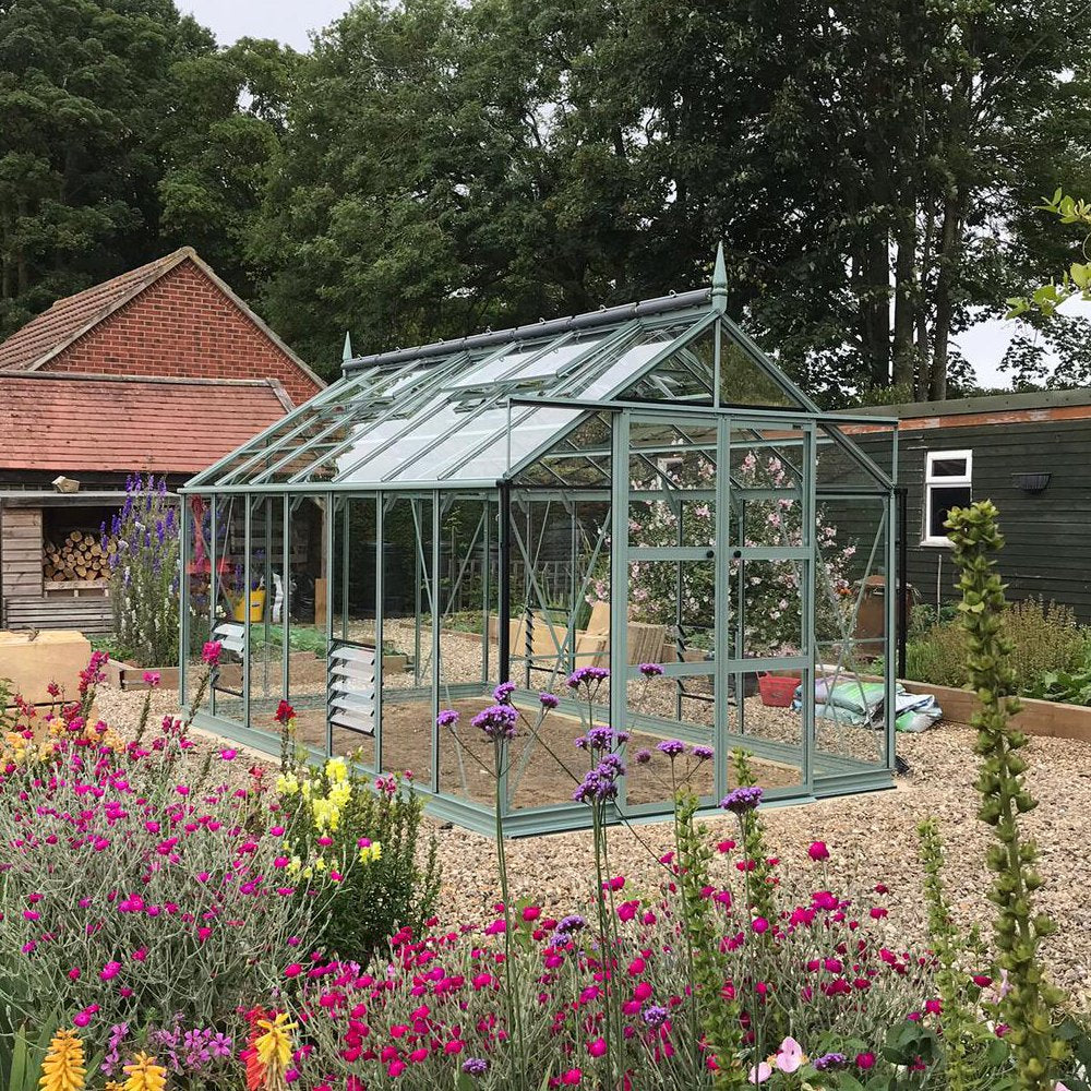 Rhino Greenhouse stands on gravel, surrounded by colorful flowering plants, with a wooden shed and trees in the background. Fiberglass panels reflect soft light, creating a serene gardening space.