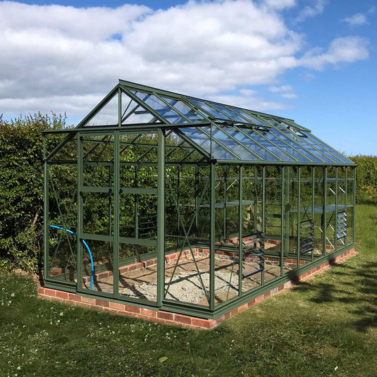 A green metal-framed Rhino Greenhouse with transparent glass panels stands in a grassy area, surrounded by hedges and under a partly cloudy sky.