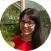 A person wearing glasses and a red shirt is smiling outdoors, standing near green foliage and a glass structure, suggesting a garden or Rhino Greenhouse environment.