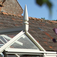 A decorative finial sits atop the glass roof of a Rhino Greenhouse, positioned against a backdrop of an old, weathered brick building with a tiled roof under a clear blue sky.