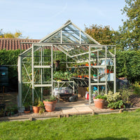 A glass Rhino Greenhouse shelters plants and gardening tools, positioned in a garden with grass and hedges, reflecting sunlight on a clear day.