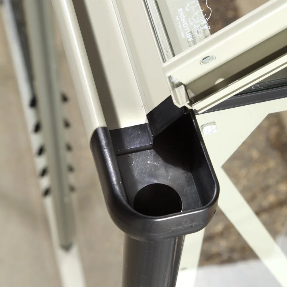 A corner of a metal and glass structure with a drainage system, featuring a RHINOTUFF label, is part of a Rhino Greenhouse support beam, surrounded by a concrete floor.