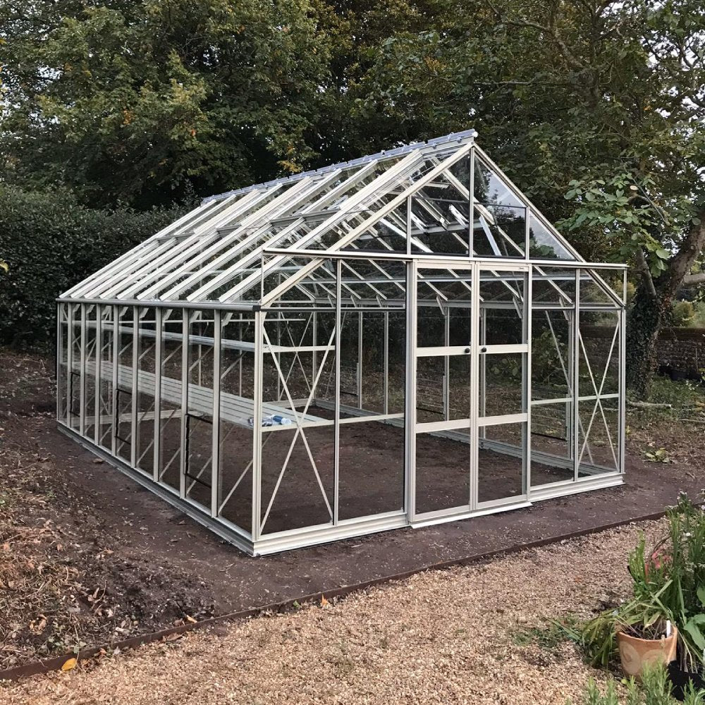 A large glass Rhino Greenhouse stands on a prepared soil foundation, surrounded by trees and greenery. The structure features a sliding door and triangular roof panels.