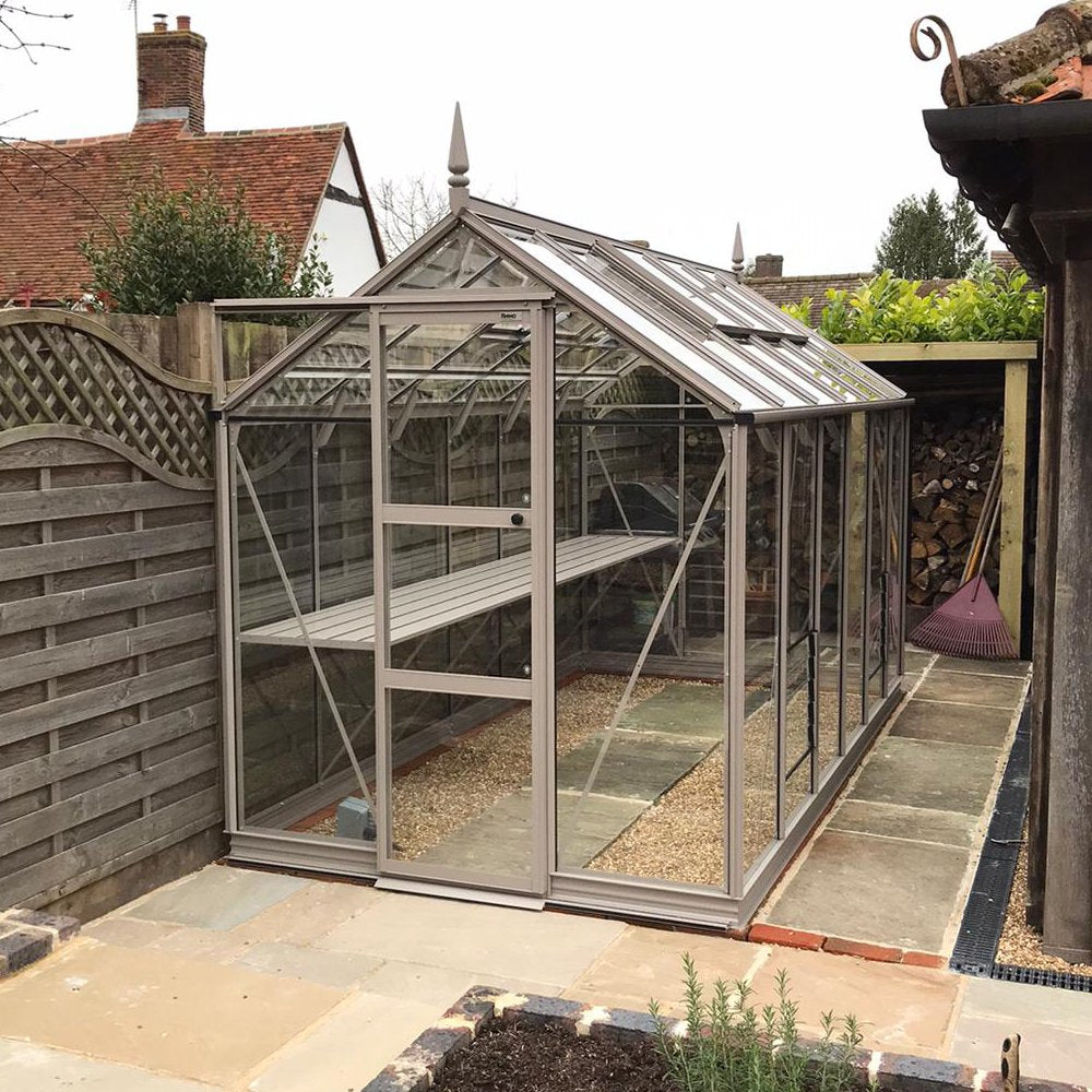 A glass Rhino Greenhouse stands in a neatly arranged backyard, bordered by a wooden fence and adjacent to a woodpile. Stone and gravel pathways surround the structure, enhancing the garden setting.