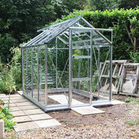A glass Rhino Greenhouse stands on a paved area in a garden, surrounded by lush greenery and wooden pallets.