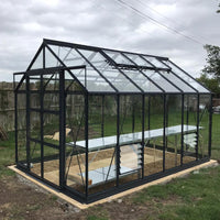 A glass and metal Rhino Greenhouse stands on a wooden base, with vented windows and internal shelves, situated in a grassy area enclosed by a wooden fence under a cloudy sky.