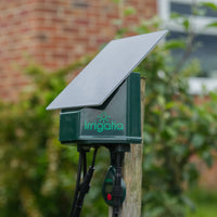 Solar-powered irrigation device mounted on a wooden post with Irrigatia label, surrounded by garden greenery and a blurred brick wall in the background.