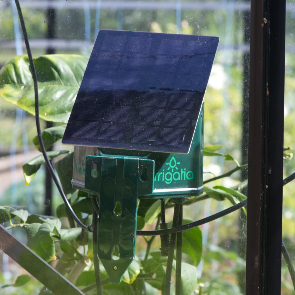 A solar panel device, labeled irrigatia, is mounted on a green frame, surrounded by lush plant foliage, inside a glass structure.