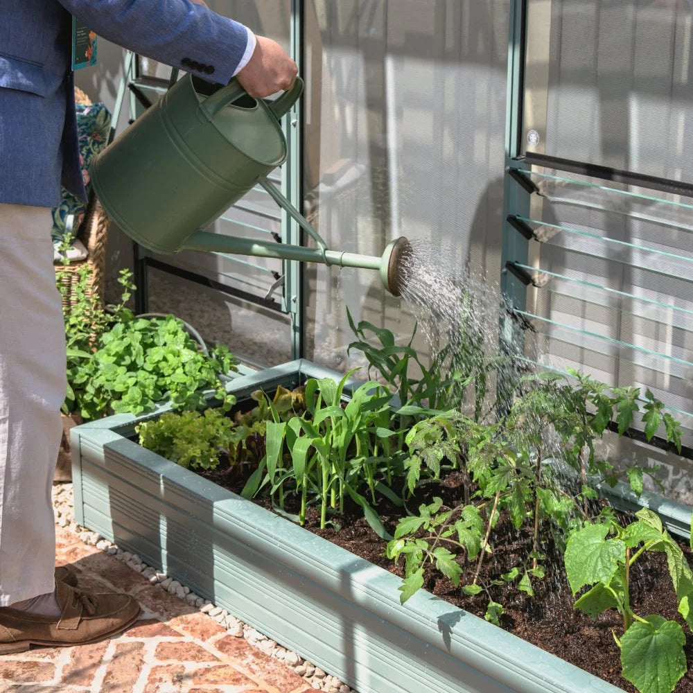 A person waters young plants in a Rhino metal raised garden bed inside a Rhino Greenhouse. The surrounding environment includes green foliage and sunlight filtering through the Rhino Greenhouse structure.