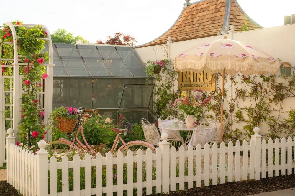 A vintage pink bicycle with a flower basket is parked by a glass Rhino Greenhouse. Nearby, a table with chairs and an umbrella sits by a white picket fence. Sign reads RHINO Rhino Greenhouses/direct.