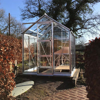 A glass and metal Rhino Greenhouse stands on a paved patio surrounded by dry hedges, under a clear sky with tall trees in the background.