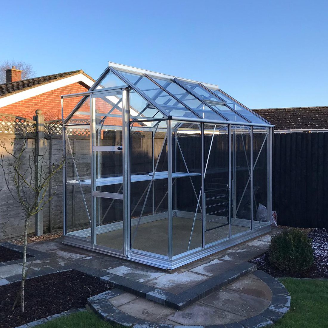 A glass Rhino Greenhouse with aluminum framing stands on a paved patio. It features interior shelving, surrounded by a garden with neatly trimmed grass and bordered by wooden fencing.