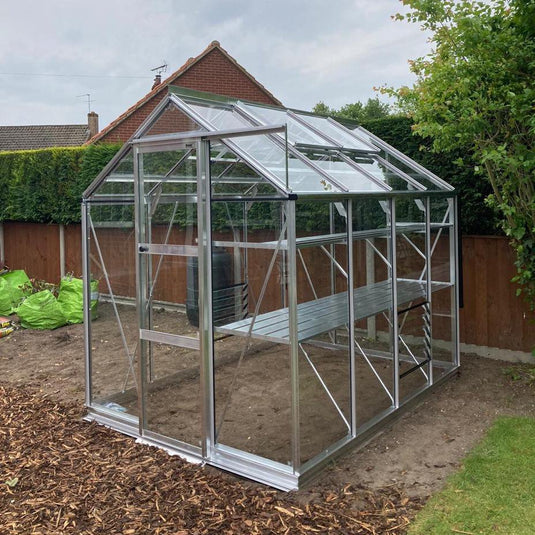 A glass-paneled Rhino Greenhouse stands in a backyard, surrounded by mulch, soil, and green hedges. A house with a sloped roof is visible in the background.