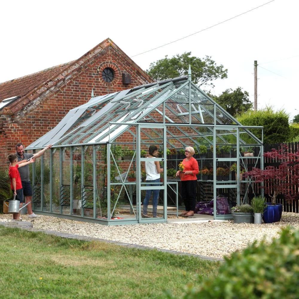 Rhino Greenhouse with clear panels houses three people engaging in gardening activities; surrounded by plants and gravel, adjacent to a rustic brick building and lush greenery.