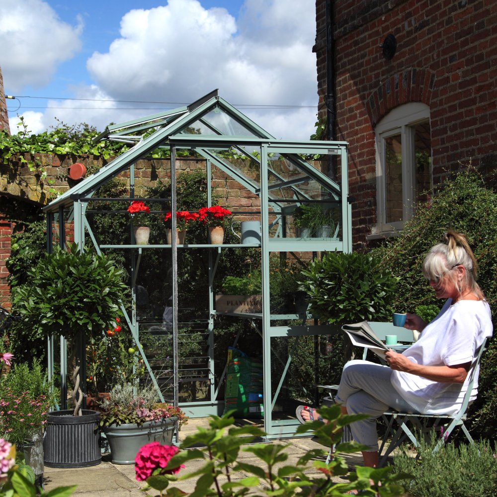 Rhino Greenhouse with potted plants inside, surrounded by brick walls and foliage. A person sits nearby in a chair, reading a newspaper and holding a blue cup, under a sunny blue sky.