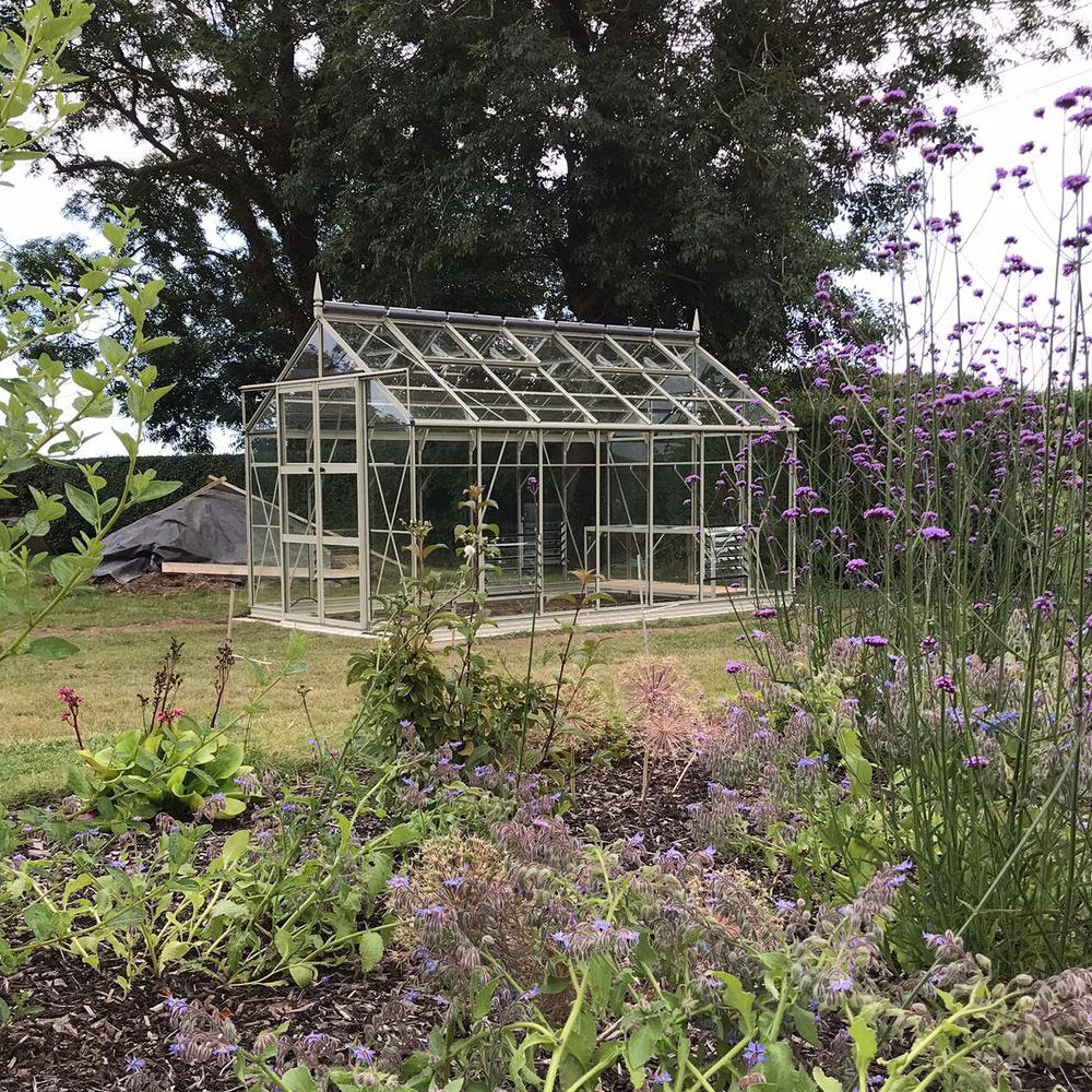 A glass Rhino Greenhouse sits on a grassy lawn surrounded by colorful flowering plants and trees. The sky is partly visible through the branches above.
