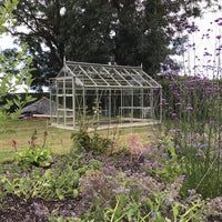 A glass Rhino Greenhouse sits on a grassy lawn surrounded by colorful flowering plants and trees. The sky is partly visible through the branches above.