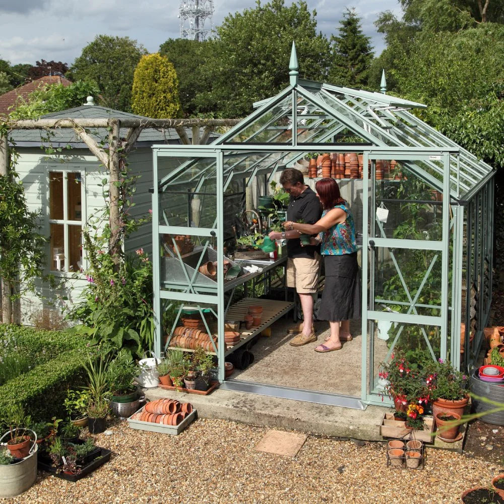 A glass Rhino Greenhouse houses two people arranging plants and pots on shelves. Surrounding it is a lush garden with various potted plants and a wooden shed.