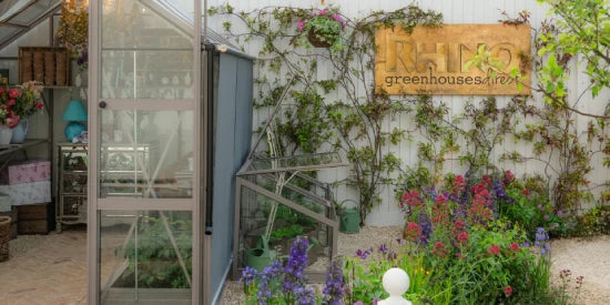 A Rhino Greenhouse entrance stands open, revealing gardening supplies, under a vine-covered wall with a Rhino Rhino Greenhouses direct sign. Surrounding vibrant flowers add color to the scene.