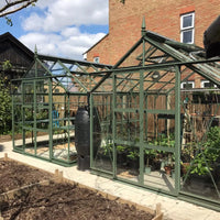 Two green metal-framed glass Rhino Greenhouses housing various plants sit in a backyard garden. A black water barrel stands beside the Rhino Greenhouse, surrounded by a wooden fence and neighboring houses.