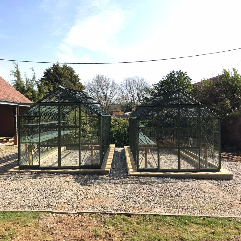 Two glass Rhino Greenhouses rest on gravel foundations, surrounded by trees and buildings under a clear sky.