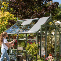 A Rhino Greenhouse with open roof panels is being adjusted by a person using a pole. Inside, there are potted plants and gardening accessories. Surrounding the structure are lush trees and shrubs.