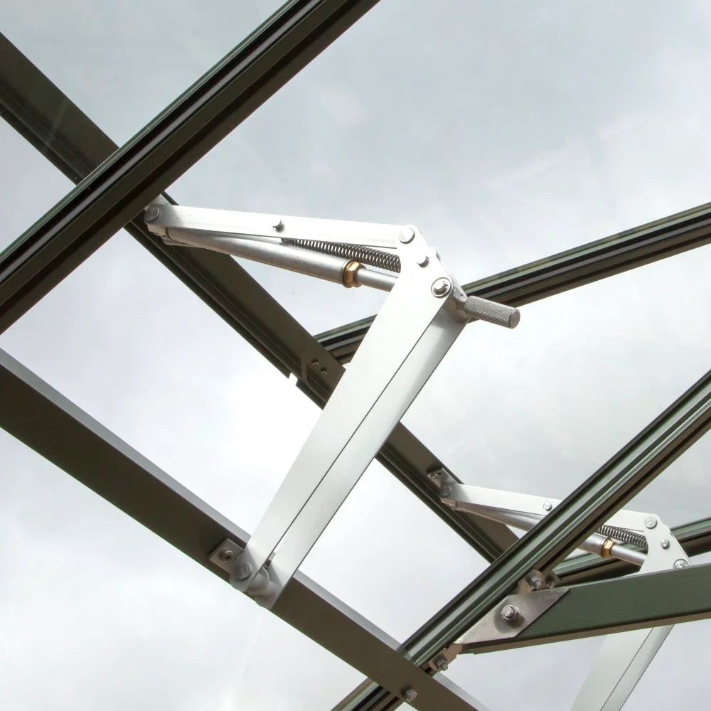 A metal window hinge mechanism opens a glass panel in a Rhino Greenhouse. The overcast sky is visible through the glass panes, emphasizing the outdoor environment.