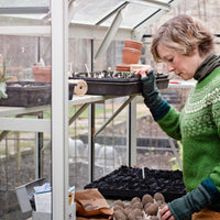 Person examining seedlings in a Rhino Greenhouse, wearing a green sweater and gloves. Surrounding them are trays of plants, pots, and gardening tools, creating a nurturing gardening environment.